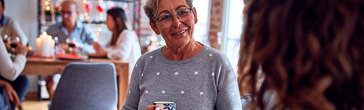 Frau mit Kaffeetasse in der Hand im Gespräch, im Hintergrund weitere Menschen am Tisch